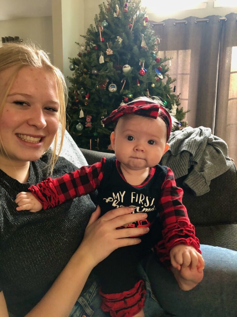 Basia, a teenage girl from Poland, is holding a baby in a Christmas outfit, with a large Christmas tree standing behind them.