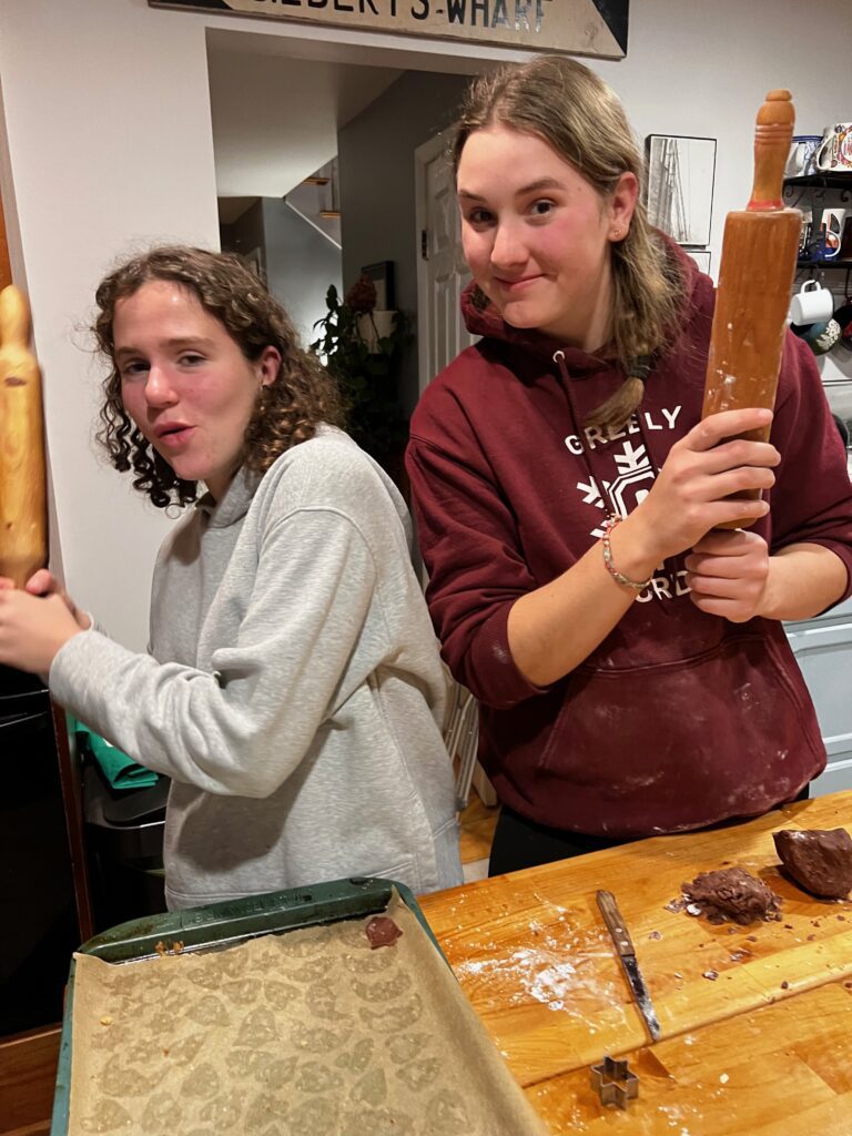 Two teenage girls, Elena and Nela, hold rolling pins, pretending they're swords, over the kitchen counter as they bake cookies. Faith is hosting them this year. 