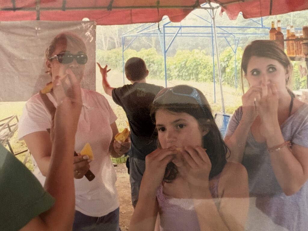 Two adult women and a young girl eat fruit underneath a tent in a tropical location.